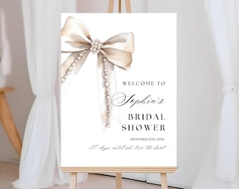 May include: A white rectangular sign featuring a watercolour illustration of a beige ribbon bow adorned with pearls. The text reads: Welcome to Sophia's Bridal Shower, September 23th, 20XX, and 30 days until she ties the knot in black lettering.