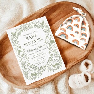 May include: A wooden tray holds a baby shower invitation, a rainbow-patterned hat, and white baby shoes. The invitation features a floral border and text. The hat has a repeating rainbow design.