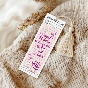 May include: A pink library card-style bookmark with the text "Consent? Oh baby, it's signed, sealed, and moaned." The bookmark has a tassel and a lip graphic. The background is a cream-colored knitted blanket.