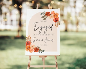 May include: A rectangular engagement sign featuring a beige arch, watercolor red and orange floral accents, and black text that reads: WE ARE Engaged LET'S CELEBRATE! Sara & James 09.25.2024. The sign is displayed on a wooden easel.