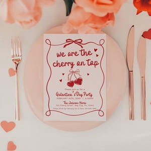 May include: A Valentine's Day party invitation with the text "we are the cherry on top" in red script. The invitation is on a pink plate, with a fork and knives. Scattered heart-shaped confetti and flowers complete the romantic setting.