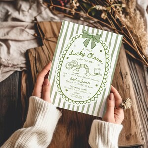May include: A green and white baby shower invitation with the text "A Little Lucky Charm Is On The Way." The invitation features a shamrock, rainbow, and leprechaun hat design. The invitation is held by a person.