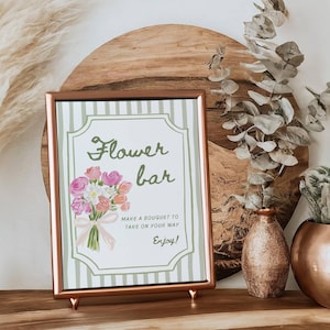 May include: A framed sign with the words "Flower bar" and a watercolor bouquet illustration. The sign is in a copper-colored frame and is propped up on a wooden surface. Decorative vases and plants are in the background.