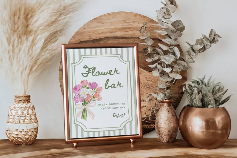 May include: A framed sign with the words "Flower bar" and a watercolor bouquet illustration. The sign is in a copper-colored frame and is propped up on a wooden surface. Decorative vases and plants are in the background.