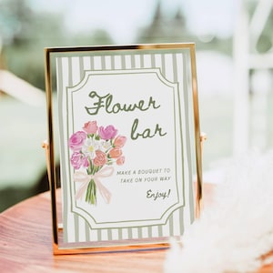 May include: A gold-framed sign with the words "Flower bar" in elegant script, featuring a watercolor bouquet of pink and peach flowers. The sign includes the text "Make a bouquet to take on your way. Enjoy!"