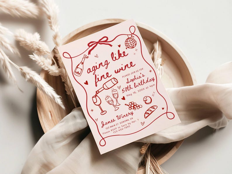 May include: A light pink invitation card with the text "aging like fine wine" and decorative wine-themed illustrations. The card is for Sophia's 50th birthday, with the date and time included. The card is placed on a wooden tray with decorative elements.