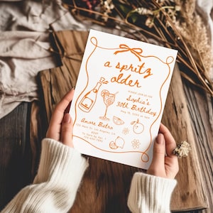 May include: A white invitation card with orange text and illustrations is held by a person. The card reads "a spritz older" and invites guests to Sophia's 30th birthday. Illustrations include a champagne bottle, cocktail, and fruit.
