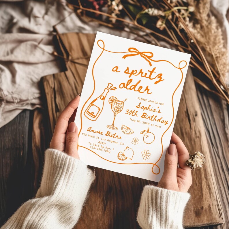 May include: A white invitation card with orange text and illustrations is held by a person. The card reads "a spritz older" and invites guests to Sophia's 30th birthday. Illustrations include a champagne bottle, cocktail, and fruit.