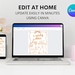 May include: A laptop, smartphone, and a pink mug are on a white surface. The laptop and smartphone display a birthday invitation design with the text "a spritz older". The invitation is orange on a white background. The text "EDIT AT HOME" is at the top.