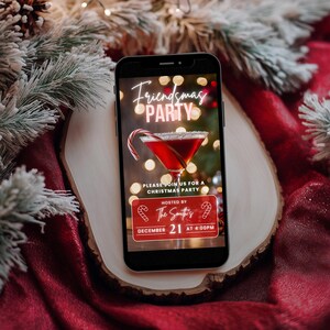 May include: A smartphone displaying a digital invitation for a Friendsmas party. The invitation features a festive red cocktail with a candy cane garnish and the text "Friendsmas Party" in neon pink letters. The text also includes the date and time of the party: December 21 at 4:00 PM.