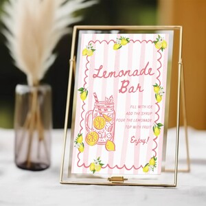 May include: A framed sign with a pink and white striped background, featuring the words "Lemonade Bar" in a decorative font. The sign includes a drawing of a lemonade drink and instructions. Yellow lemons and green leaves decorate the edges.