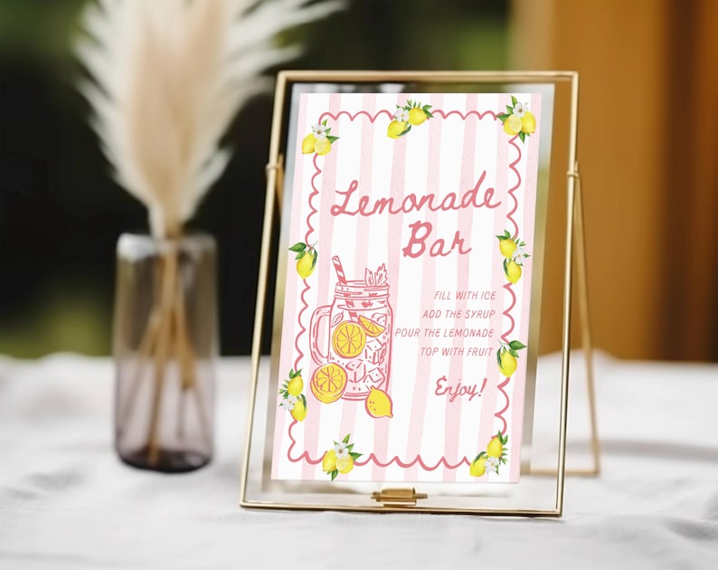 May include: A framed sign with a pink and white striped background, featuring the words "Lemonade Bar" in a decorative font. The sign includes a drawing of a lemonade drink and instructions. Yellow lemons and green leaves decorate the edges.