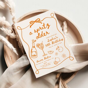 May include: A cream-colored invitation with orange text and illustrations. The text reads "a spritz older" and announces Sophia's 30th birthday. The design includes a bottle, cocktail, and fruit illustrations, with the event details and RSVP information.