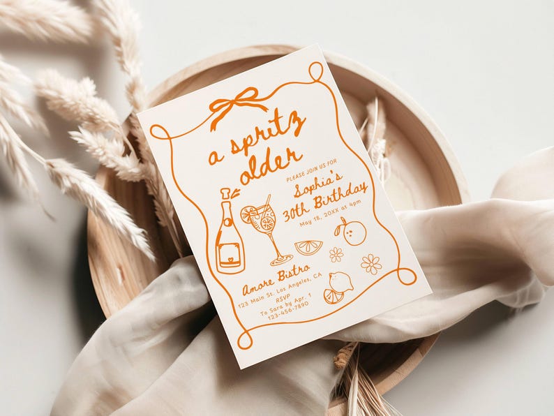 May include: A cream-colored invitation with orange text and illustrations. The text reads "a spritz older" and announces Sophia's 30th birthday. The design includes a bottle, cocktail, and fruit illustrations, with the event details and RSVP information.