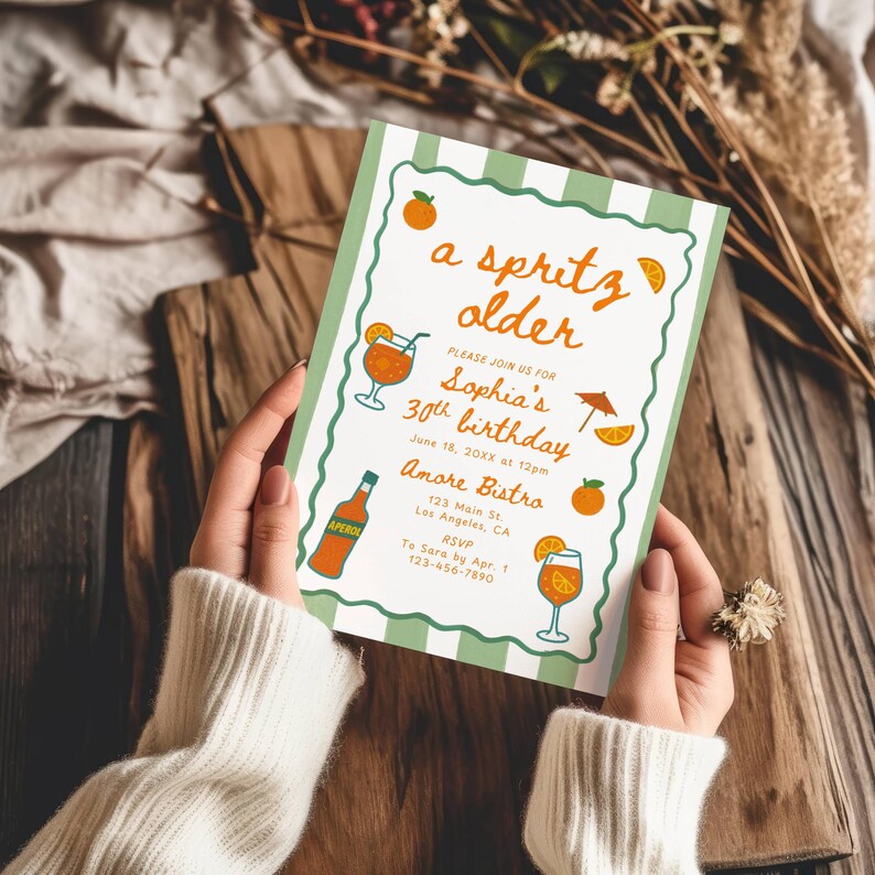 Op de afbeelding: Een groen en wit gestreepte uitnodiging met oranje accenten, vastgehouden door een persoon. De uitnodiging bevat de tekst "a spritz older" en illustraties van cocktails, sinaasappels en een fles Aperol, voor een 30e verjaardag.