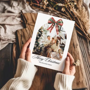 May include: A Christmas card with a red and green plaid bow on top of a photo of a family standing in front of a Christmas tree. The card reads "Merry Christmas" and "The Williams Family".