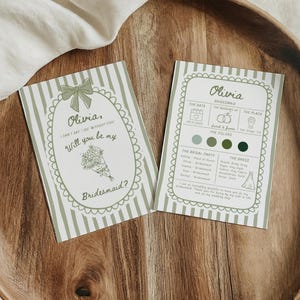May include: Two invitation cards on a wooden tray. The cards are light green with white stripes and decorative borders. One card reads "Olivia, Will you be my Bridesmaid?" The other card has wedding details. The tray is made of wood.
