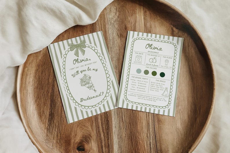 May include: Two invitation cards on a wooden tray. The cards are light green with white stripes and decorative borders. One card reads "Olivia, Will you be my Bridesmaid?" The other card has wedding details. The tray is made of wood.