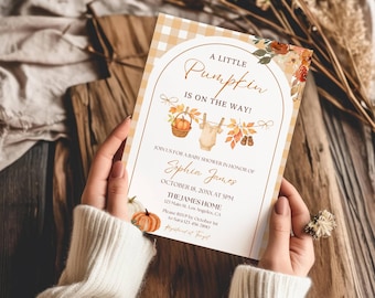 May include: A rectangular baby shower invitation card featuring a yellow and white gingham border. The text reads A LITTLE PUMPKIN IS ON THE WAY, with illustrations of a pumpkin in a basket, a baby onesie, and autumn leaves on a clothesline.
