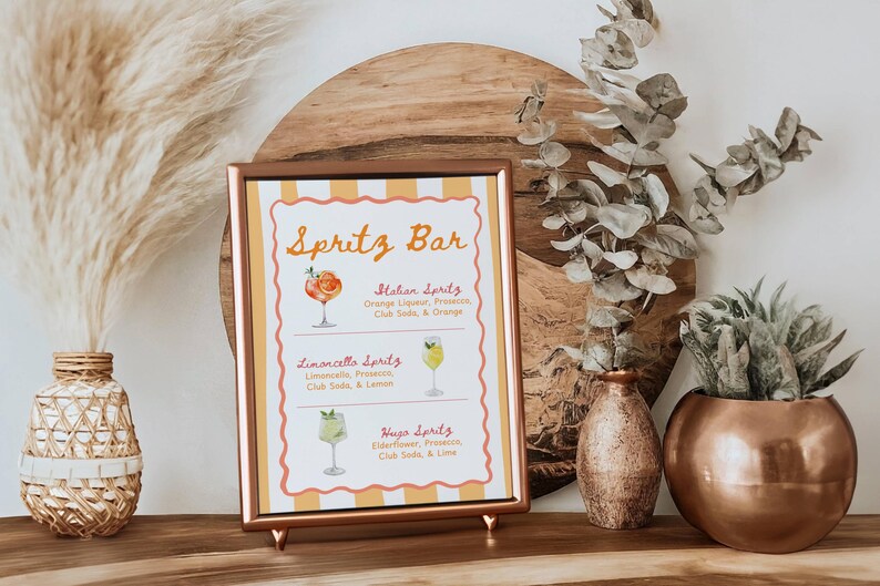May include: A framed sign with the text "Spritz Bar" and drink recipes. The sign is in a copper-colored frame and is propped up on a wooden surface. Decorative vases and plants are in the background.