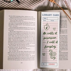 May include: A bookmark with a library card design, featuring the text "He calls it possession, I call it foreplay." Includes a heart, a lock, and a snake illustration. The bookmark is set on an open book.