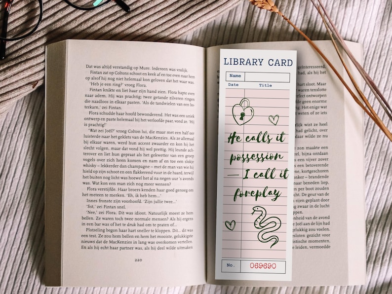 May include: A bookmark with a library card design, featuring the text "He calls it possession, I call it foreplay." Includes a heart, a lock, and a snake illustration. The bookmark is set on an open book.