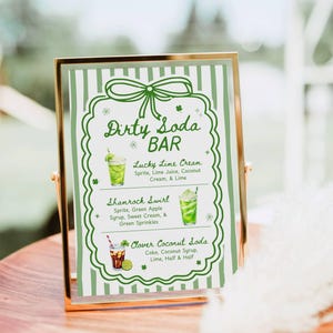 May include: A framed sign with a gold border displays a "Dirty Soda Bar" menu. The menu features three drink options: Lucky Lime Cream, Shamrock Swirl, and Clover Coconut Soda. The sign has a green and white striped background with green decorative elements.