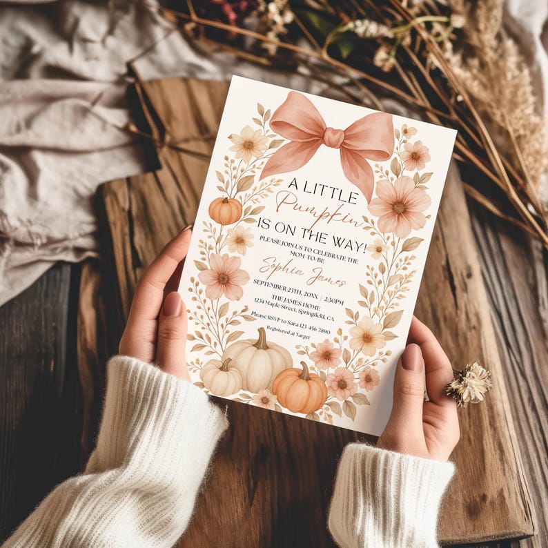 May include: A baby shower invitation with a fall theme. The card features a peach bow, pumpkins, and floral accents. Text reads "A Little Pumpkin is on the Way!" and includes event details. The invitation is held by a person.