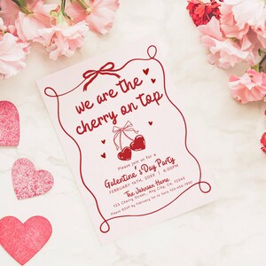May include: A pink Galentine's Day party invitation with the text "we are the cherry on top" in red script. The invitation features a cherry illustration and heart accents. Pink paper hearts and flowers surround the invitation.