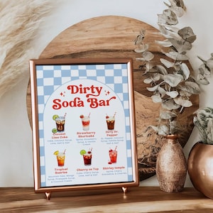 May include: A framed "Dirty Soda Bar" sign with a light blue and white checkered background. The sign features illustrations of six soda drinks with their ingredients listed below. The sign is displayed on a wooden surface.