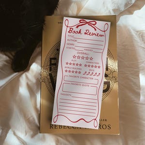 Puede incluir: Una tarjeta rectangular blanca de reseña de libros con texto y gráficos en tinta roja. La tarjeta incluye campos para título, autor, género, fechas y valoraciones de trama, personajes, construcción del mundo y picante, además de espacios para citas.