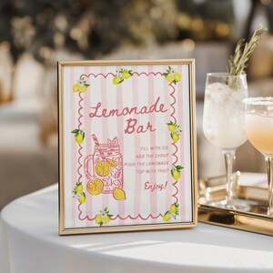 May include: A framed sign with a gold border, featuring a pink and white striped background. The sign reads "Lemonade Bar" in cursive, with a drawing of a lemonade drink and instructions. Yellow lemons and green leaves decorate the edges.