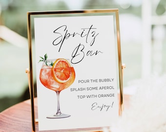 May include: A gold-framed sign with the text "Spritz Bar" in black cursive font. An illustration of a glass of Aperol Spritz with an orange slice is shown. The instructions for making the drink are: "Pour the bubbly, splash some Aperol, top with orange, Enjoy!"