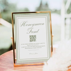 May include: A rectangular sign in a gold metal frame featuring the text Honeymoon Fund, a QR code, and a poem about cash gifts. The sign has a white background with sage green striped borders and the names Olivia & James printed at the bottom.