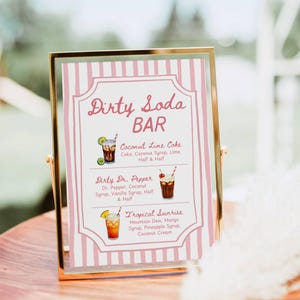 May include: A gold-framed sign with a pink and white striped background. The sign reads "Dirty Soda BAR" and lists three soda drink recipes: Coconut Lime Coke, Dirty Dr. Pepper, and Tropical Sunrise. The sign is displayed on a wooden surface.