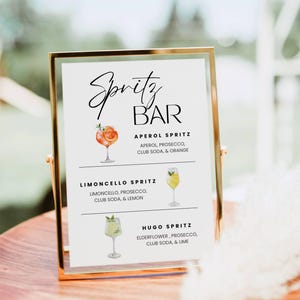 Könnte beinhalten: Ein goldgerahmtes Schild mit der Aufschrift "Spritz BAR" in eleganter Schrift. Das Schild listet drei Spritz-Cocktails auf: Aperol Spritz, Limoncello Spritz und Hugo Spritz, jeweils mit einer Aquarellillustration des Getränks und seiner Zutaten.