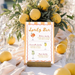 May include: A table setting with a "Spritz Bar" sign featuring three spritz cocktail recipes: Italian, Limoncello, and Hugo. The sign is surrounded by lemons and floral arrangements, creating a vibrant and inviting atmosphere.