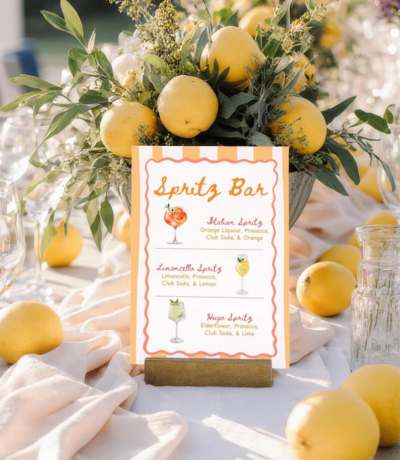 May include: A table setting with a "Spritz Bar" sign featuring three spritz cocktail recipes: Italian, Limoncello, and Hugo. The sign is surrounded by lemons and floral arrangements, creating a vibrant and inviting atmosphere.