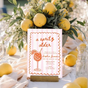 May include: A pink and white striped invitation with the words "a spritz older" in red script. The invitation features a cocktail illustration and event details. Lemons and greenery surround the invitation, creating a festive atmosphere.
