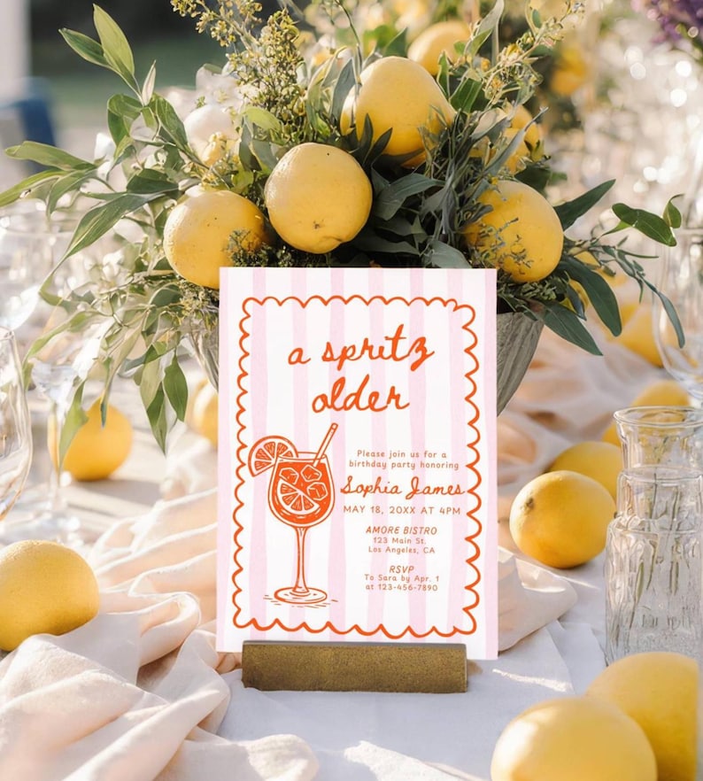 May include: A pink and white striped invitation with the words "a spritz older" in red script. The invitation features a cocktail illustration and event details. Lemons and greenery surround the invitation, creating a festive atmosphere.