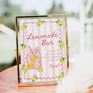 May include: A framed sign with a gold border displays "Lemonade Bar" in pink script over pink and white stripes. The sign includes instructions and a lemon illustration. The sign is on a wooden surface.