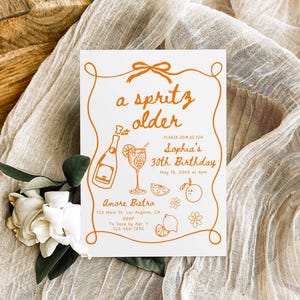 May include: A white invitation card with orange text and illustrations, featuring the phrase "a spritz older" and details for Sophia's 30th birthday. The card includes a bottle, cocktail, and fruit illustrations. The card is on a beige fabric.