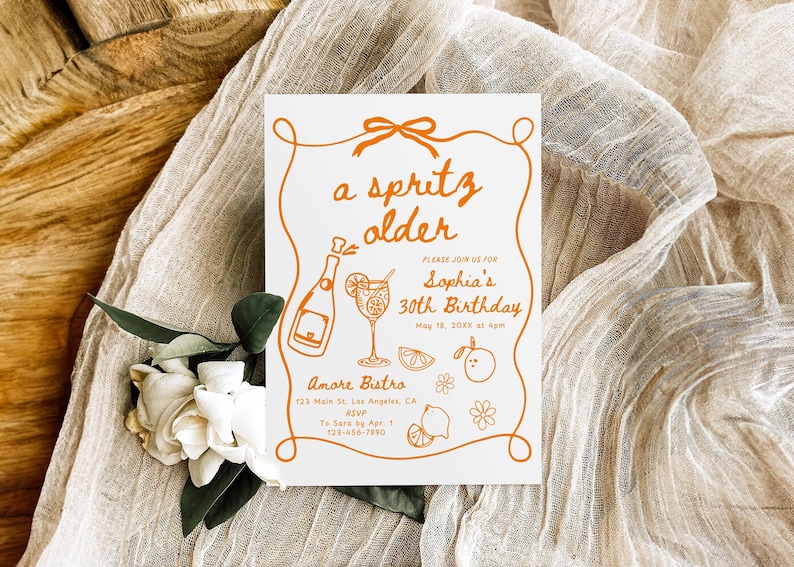 May include: A white invitation card with orange text and illustrations, featuring the phrase "a spritz older" and details for Sophia's 30th birthday. The card includes a bottle, cocktail, and fruit illustrations. The card is on a beige fabric.