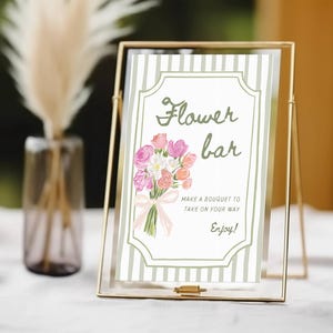 May include: A gold metal framed sign with the words "Flower bar" and a bouquet illustration. The sign includes the text "Make a bouquet to take on your way. Enjoy!" The sign is on a white surface.