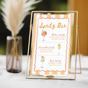 May include: A gold-framed sign with the text "Spritz Bar" in orange script. The sign lists three spritz cocktails: Italian Spritz, Limoncello Spritz, and Hugo Spritz, with watercolor illustrations of each drink.