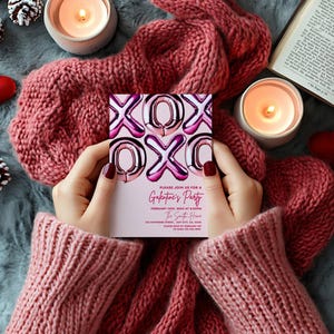 May include: A Valentine's Day party invitation with pink and silver foil balloon letters spelling XOXO is held by a person wearing a pink sweater. The invitation reads "Galentine's Party" with event details. The scene includes candles, hearts, and a cozy pink scarf.