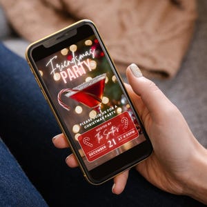 May include: A smartphone displaying a digital invitation for a Friendsmas party. The invitation features a red cocktail with a candy cane garnish, a festive background, and the text "Friendsmas Party", "Please join us for a Christmas party", "Hosted by The Smiths", "December 21 at 4:00PM".