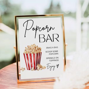 May include: A gold-framed sign with the words "Popcorn BAR" in elegant script. Below, a red and white striped popcorn container is filled with popcorn. The sign includes instructions: "Snag a bag, scoop some popcorn, sprinkle on toppings, enjoy."