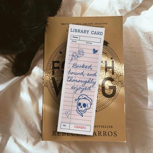 May include: A pink library card bookmark with blue floral and skull illustrations, and the text "Booked, bound, and thoroughly enjoyed." The bookmark is on top of a gold book with the title "Fourth Wing". A black cat is in the background.