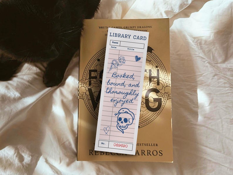 May include: A pink library card bookmark with blue floral and skull illustrations, and the text "Booked, bound, and thoroughly enjoyed." The bookmark is on top of a gold book with the title "Fourth Wing". A black cat is in the background.